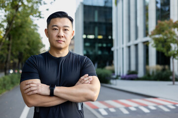 Portrait of a serious young Asian sports trainer man standing on a city street with his arms...