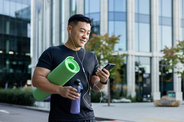 A young Asian sportsman man is standing on a city street in headphones, holding a phone, a yoga...