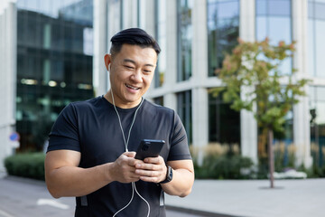 A young man, an Asian sportsman, is standing on a city street in headphones, holding a phone in his hands, making a call. dials messages, chats - obrazy, fototapety, plakaty