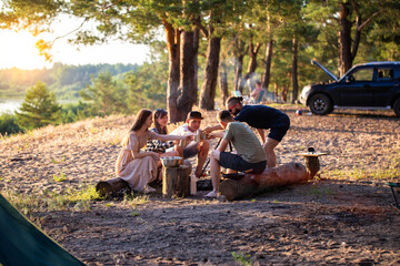 A company of friends of girls and guys is resting in the summer in a picturesque place in the forest on the banks of the river against the backdrop of sunset. Picnic in nature, tourism. Celebrating a - obrazy, fototapety, plakaty