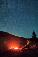 Night camping in mountains under starry sky. Woman sitting on grass near campfire, admiring...