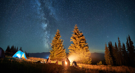 Night camping in mountains under starry sky. Group of people tourists having a rest near campsite, burning campfire and illuminated tent. Concept of tourism, hiking and adventure. - obrazy, fototapety, plakaty