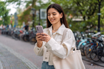 A beautiful young multiracial woman in a casual white jacket using her phone on a European street with bicycles in the background