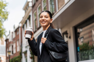 Stressless multiracial young businesswoman with a cheerful expression, in the street, holding a thermo cup of coffee