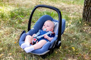 A baby boy with a pacifier in his mouth lies in a portable infant carrier on the grass in nature in the summer. Close-up