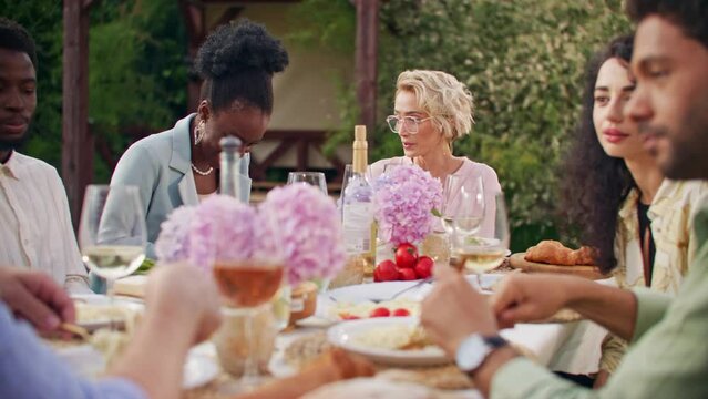 Celebration, friendship and holidays concept. Group of several multiethnic young men and women are sitting at table outdoors at home backyard, eating, communicating and clinking champagne glasses.
