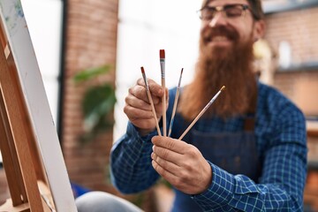 Young redhead man artist smiling confident holding paintbrushes at art studio