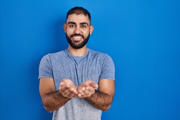 Naklejka premium Middle east man with beard standing over blue background smiling with hands palms together receiving or giving gesture. hold and protection