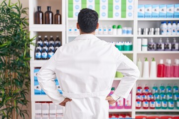 Handsome hispanic man working at pharmacy drugstore standing backwards looking away with arms on...