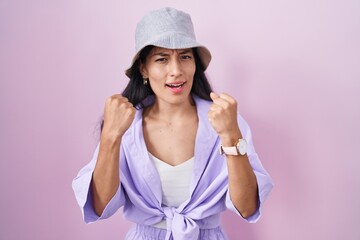 Young hispanic woman standing over pink background wearing hat angry and mad raising fists frustrated and furious while shouting with anger. rage and aggressive concept.