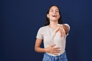 Young hispanic woman standing over blue background laughing at you, pointing finger to the camera...