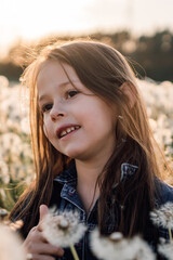 Charming little brunette girl with dark eyes stand in meadow of white dandelions at sunset and...