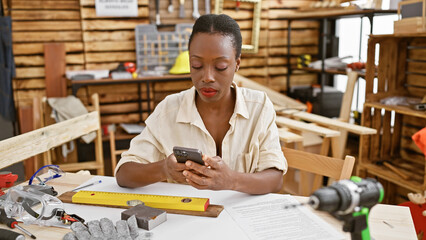 Serious african american female carpenter proficiently texting on smartphone while gracefully...