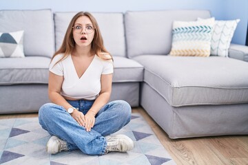 Young caucasian woman sitting on the floor at the living room afraid and shocked with surprise and amazed expression, fear and excited face.