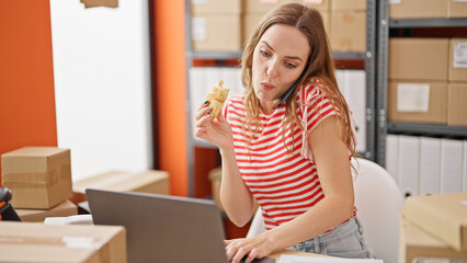 Young blonde woman ecommerce business worker eating croissant talking on smartphone working at office