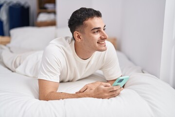 Young hispanic man using smartphone lying on bed at bedroom