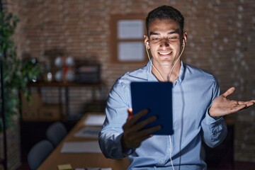 Handsome hispanic man working at the office at night pointing aside with hands open palms showing copy space, presenting advertisement smiling excited happy