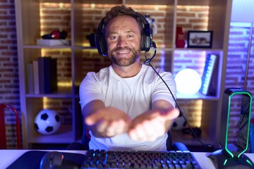 Middle age man with beard playing video games wearing headphones smiling with hands palms together receiving or giving gesture. hold and protection