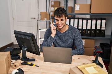 Young hispanic man ecommerce business worker using laptop talking on smartphone at office