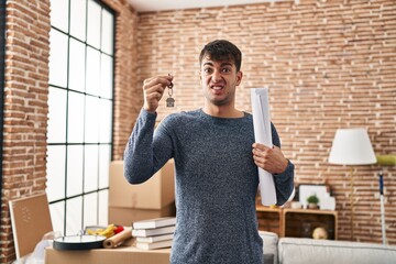 Young hispanic man holding keys of new home and blueprints clueless and confused expression. doubt...
