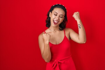 Young hispanic woman standing over red background very happy and excited doing winner gesture with arms raised, smiling and screaming for success. celebration concept.
