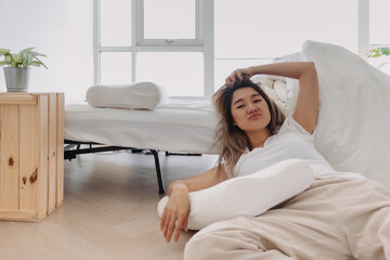 Asian Thai woman falling down from bed, sitting on floor and leaning the bed, lazy relaxing alone in home apartment, looking at camera.