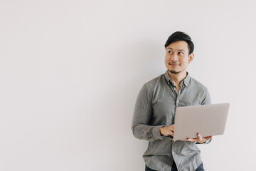 Asian man with beard wear grey shirt typing keyboard working on notebook and holding laptop, happy smiling face looking at empty space and standing isolated over white background wall. - Powered by Adobe