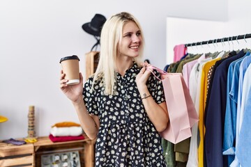 Young blonde woman smiling confident holding shopping bags and coffee at clothing store