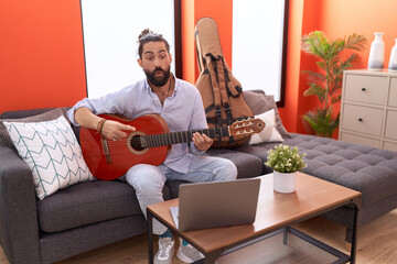 Hispanic man with beard playing classic guitar at home scared and amazed with open mouth for...