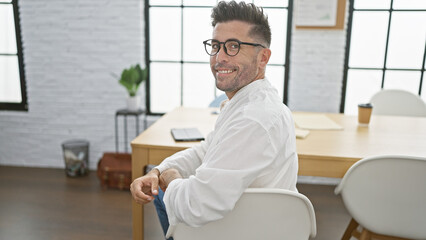 Smiling young hispanic man joyfully sits at office table, exuding confidence with business success...