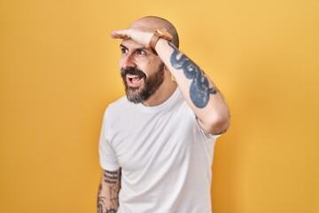 Young hispanic man with tattoos standing over yellow background very happy and smiling looking far away with hand over head. searching concept.