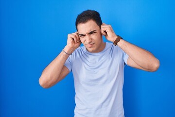 Fototapeta premium Young hispanic man standing over blue background covering ears with fingers with annoyed expression for the noise of loud music. deaf concept.