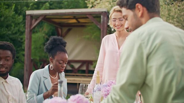 Funny mature friends laughing, sitting at table and talking with alcoholic cocktails in back yard at sunny day. Food and eating, fun at grill party outdoors. Group of diverse multi-ethnic friends.