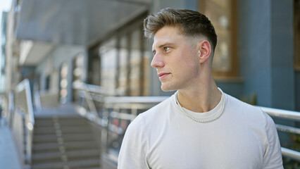 Young, handsome, caucasian man, relaxed yet exhibiting a serious expression, looking to the side whilst standing in the sunny outdoors. street fashion reveals urban, cool lifestyle.