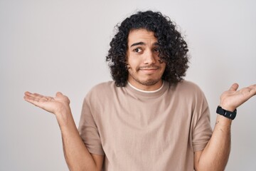 Hispanic man with curly hair standing over white background clueless and confused expression with arms and hands raised. doubt concept.