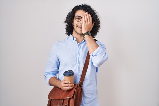 Hispanic man with curly hair drinking a cup of take away coffee covering one eye with hand, confident smile on face and surprise emotion.