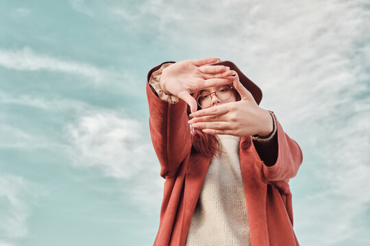 Teenage girl in a red coat and hat stands against the sky and makes a frame shot with her hands.