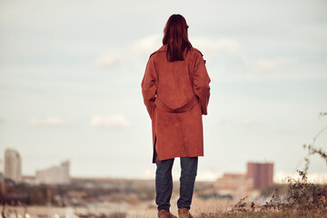 Long-haired redheaded girl in red coat stands with back to camera, hands in pockets, against blurred cityscape and sky.