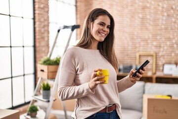 Young beautiful hispanic woman using smartphone drinking coffee at new home