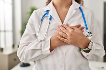 Young beautiful hispanic woman doctor standing with hands on heart at clinic