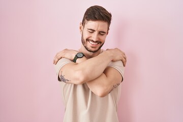 Hispanic man with beard standing over pink background hugging oneself happy and positive, smiling...