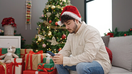 Obraz na płótnie Canvas Young hispanic man celebrating christmas using smartphone at home