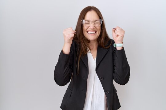 Beautiful brunette woman wearing business jacket and glasses excited for success with arms raised and eyes closed celebrating victory smiling. winner concept.