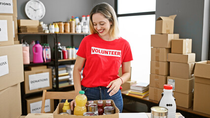 Young blonde woman volunteer holding products to donate smiling at charity center