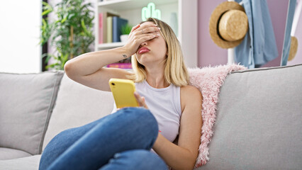 Young blonde woman using smartphone sitting on sofa looking upset at home