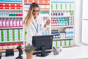 Young blonde woman pharmacist using computer holding pills tablet at pharmacy
