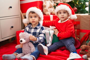 Two kids holding gift and teddy bear sitting on floor by christmas tree at home