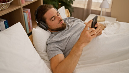 Fototapeta premium Young hispanic man using smartphone lying on the bed wearing headphones at bedroom