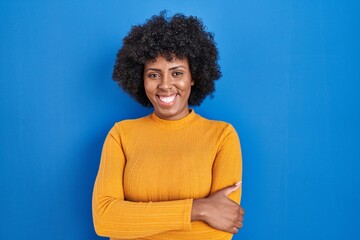 Fototapeta na wymiar Black woman with curly hair standing over blue background happy face smiling with crossed arms looking at the camera. positive person.