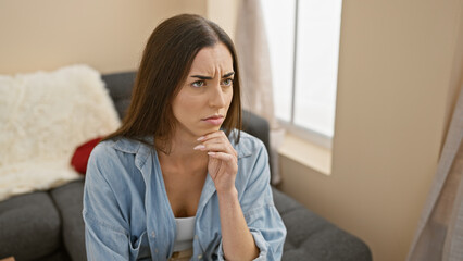Attractive young hispanic woman sitting pensively on her living room sofa, conveying a doubtful expression in a thoughtful indoor portrait
